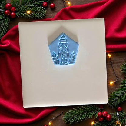 Blue nutcracker clay imprint on white tile with red fabric, pine branches, and festive lights.