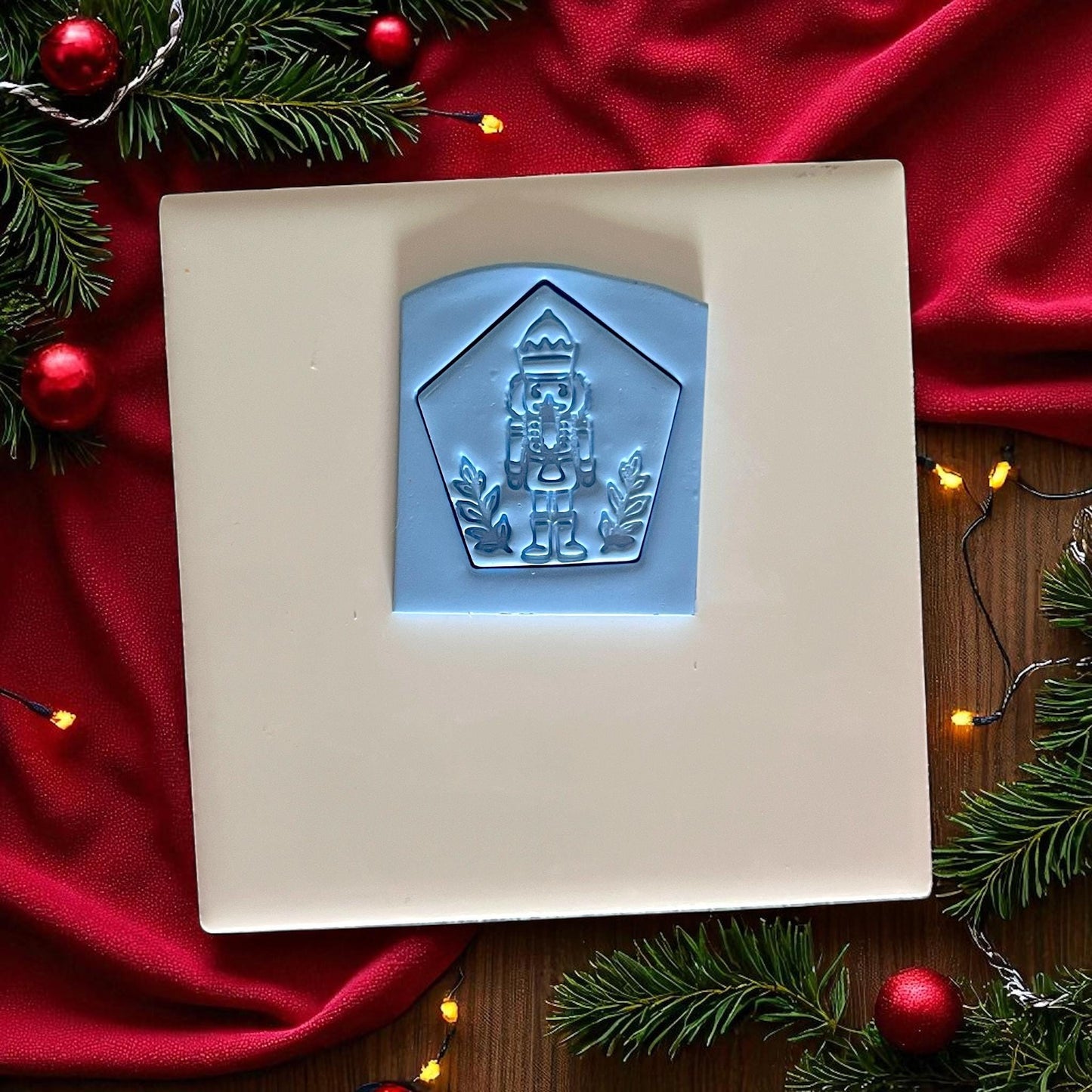 Blue clay imprint of nutcracker soldier with crown inside pentagon frame on white tile, red fabric, and Christmas decor.