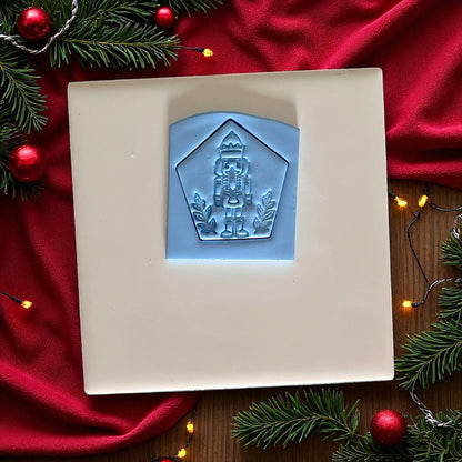 Blue clay imprint of nutcracker soldier with crown inside pentagon frame on white tile, red fabric, and Christmas decor.