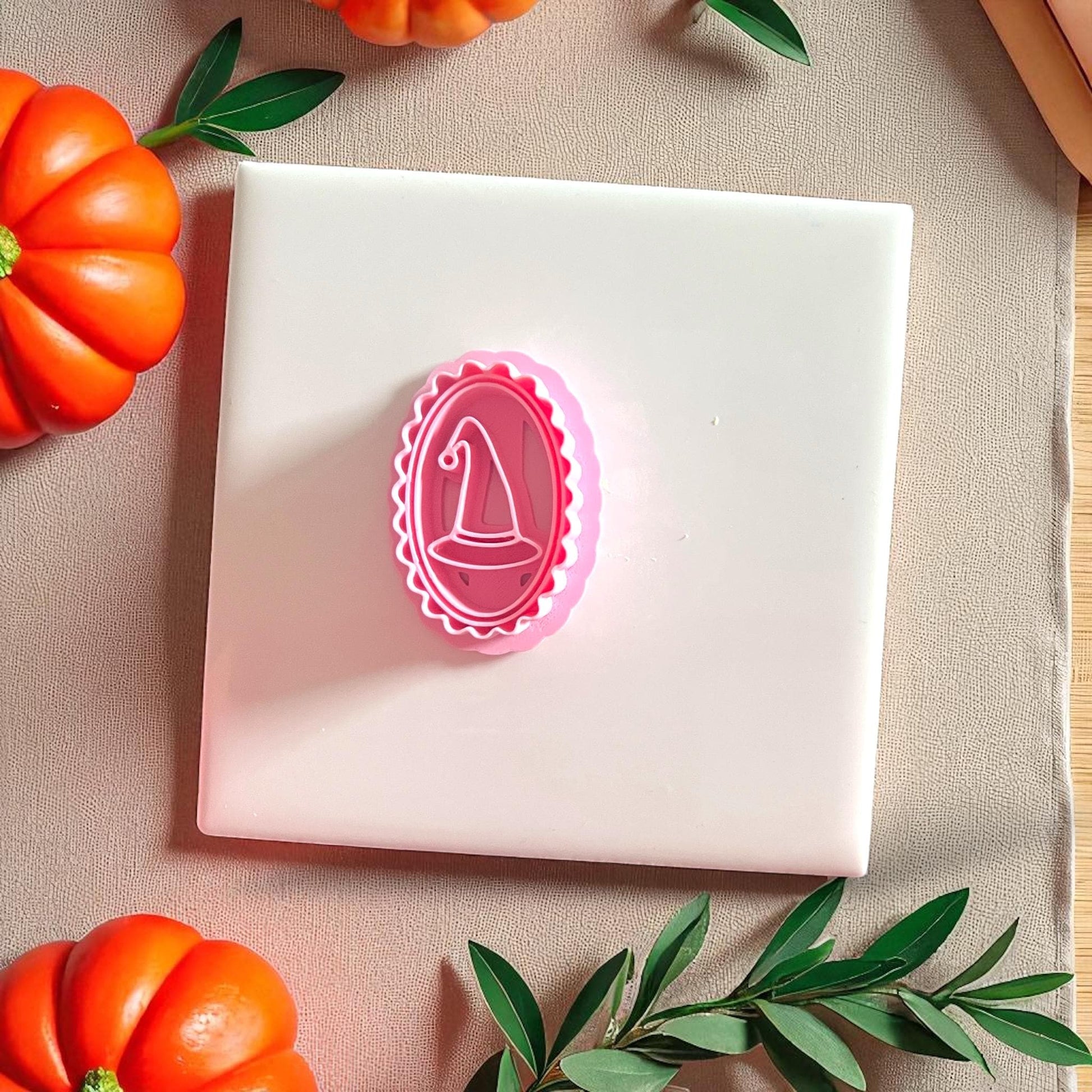 Pink oval witch hat clay cutter placed on white tile with pumpkins and greenery.