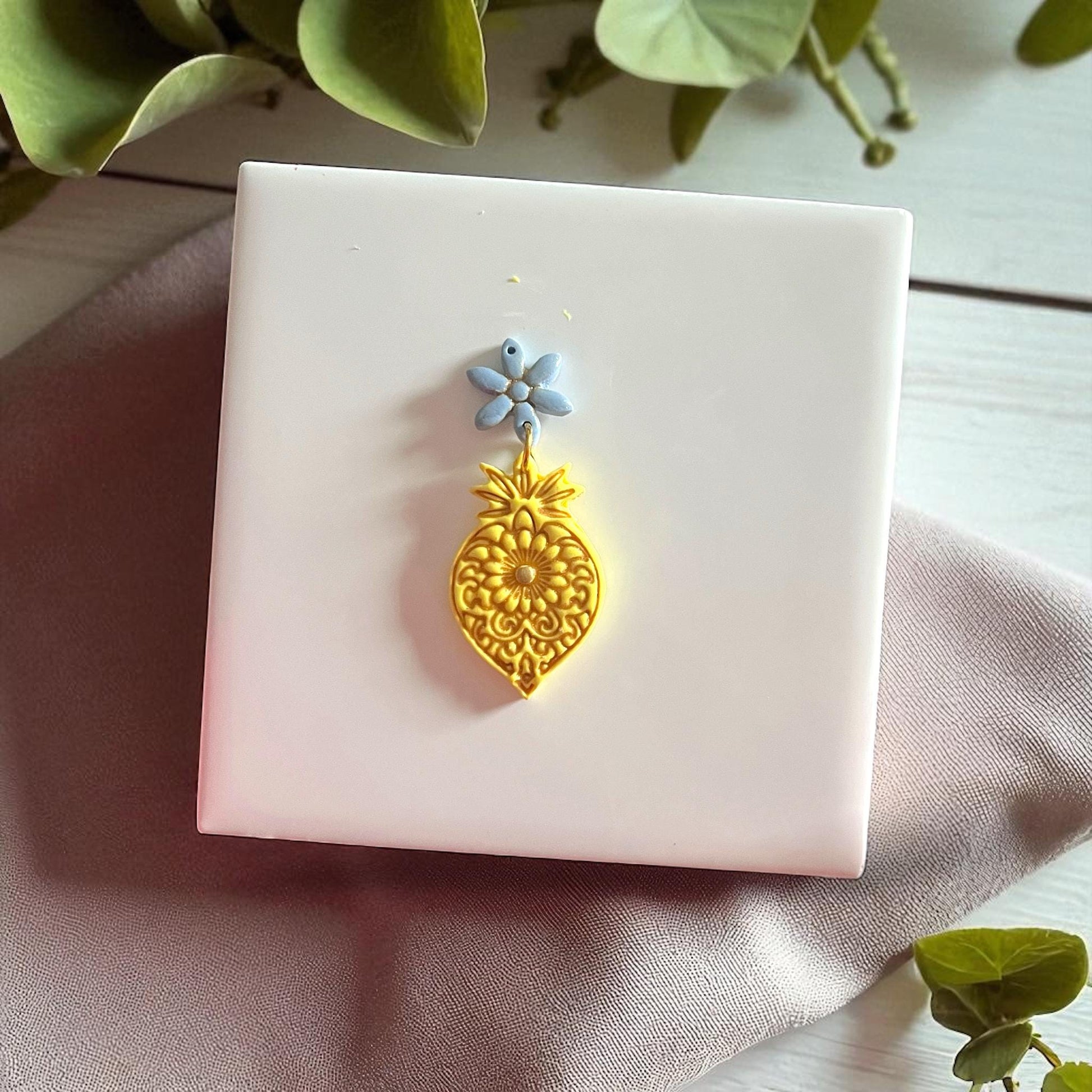 Golden mandala flower clay imprint with blue flower clay piece on white tile and fabric.