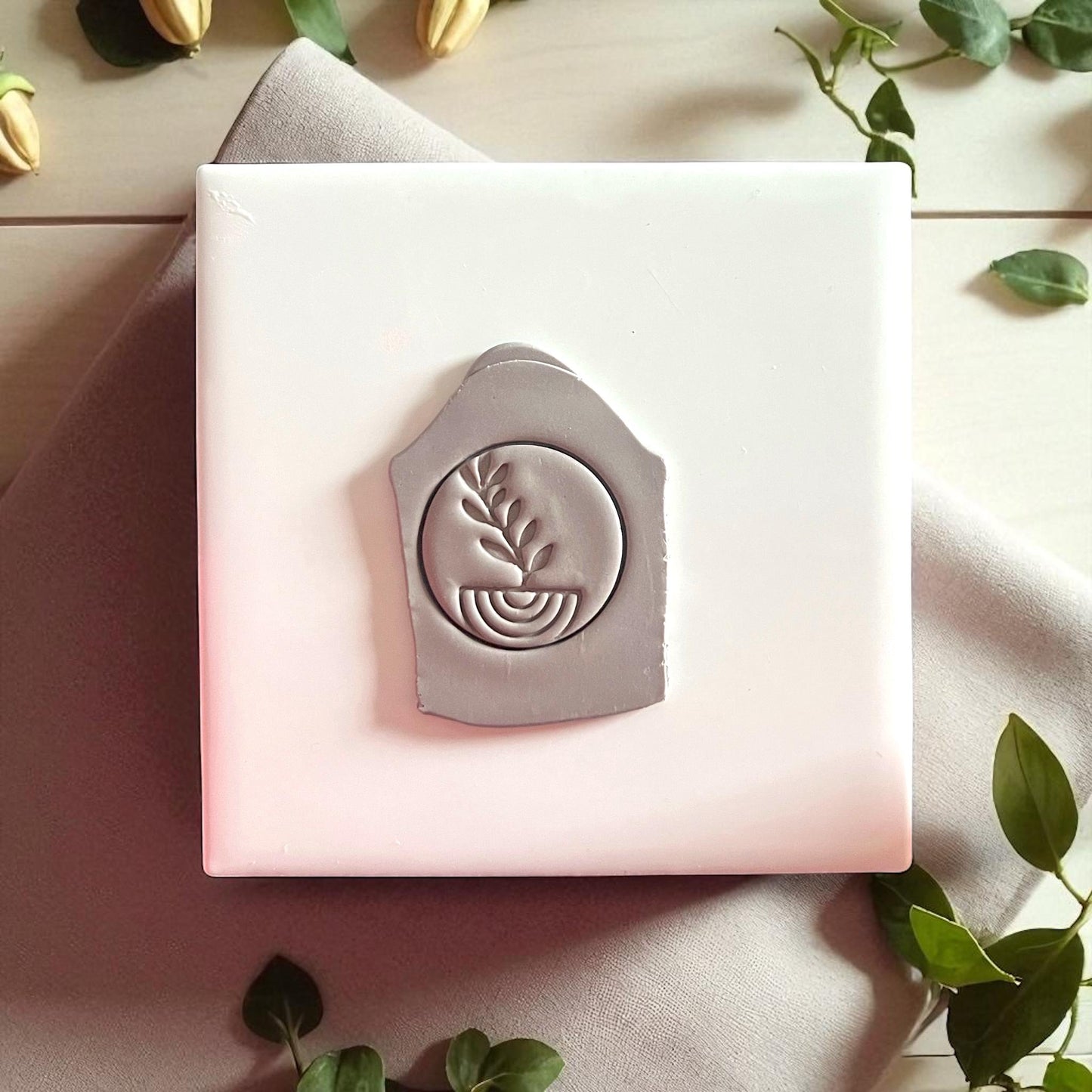 Gray clay imprint with plant and arch design on white tile with fabric and greenery.