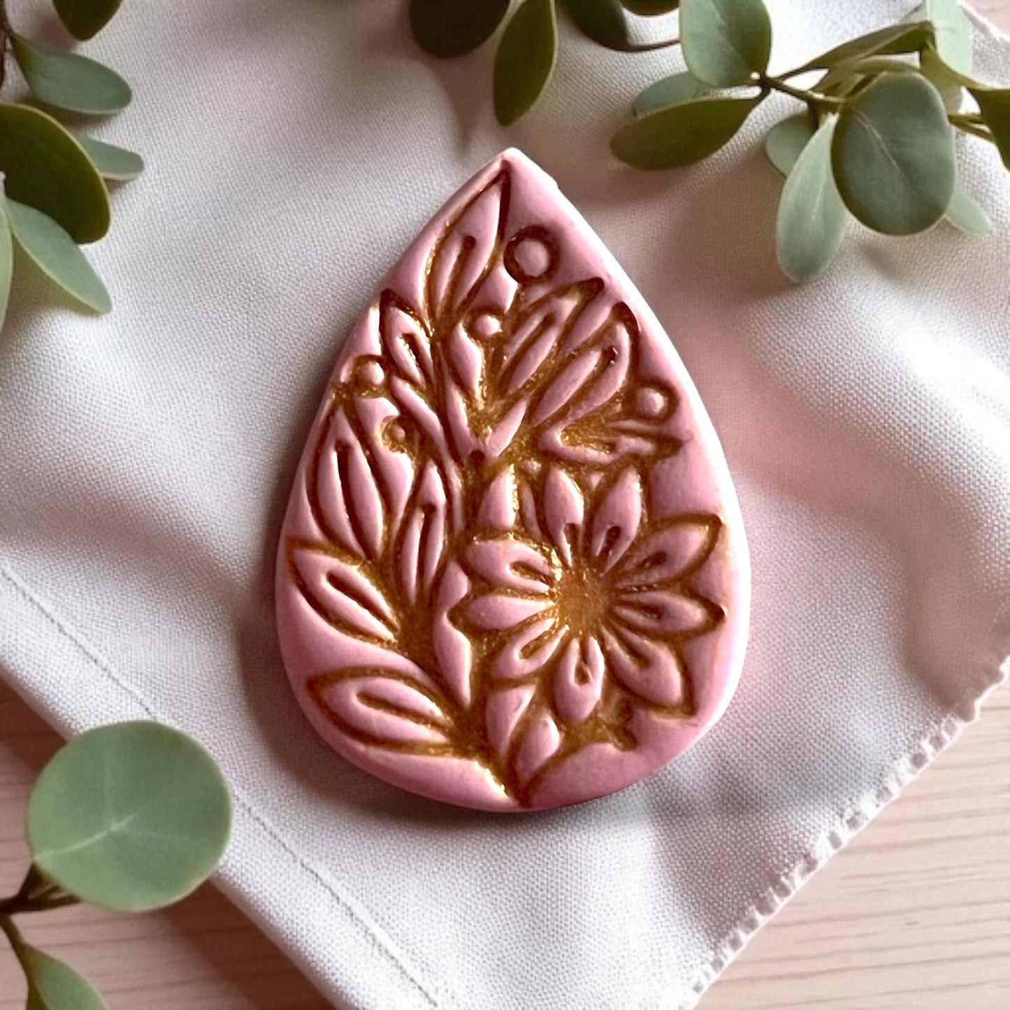 Large pink clay piece with gold painted floral teardrop design on fabric with greenery.