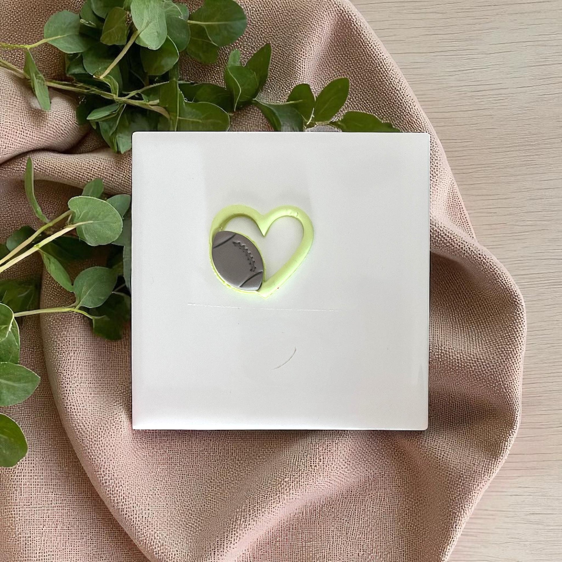 Green football heart clay piece on white tile with fabric and leafy background.