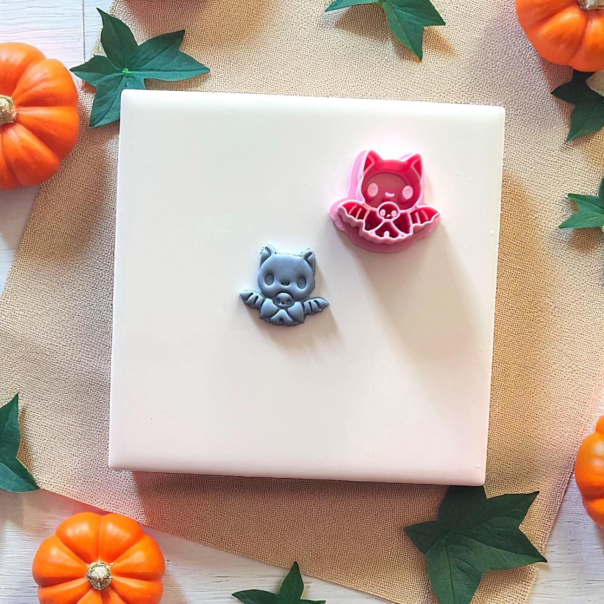 Gray baby bat clay piece beside pink bat cutter on white tile with pumpkins.