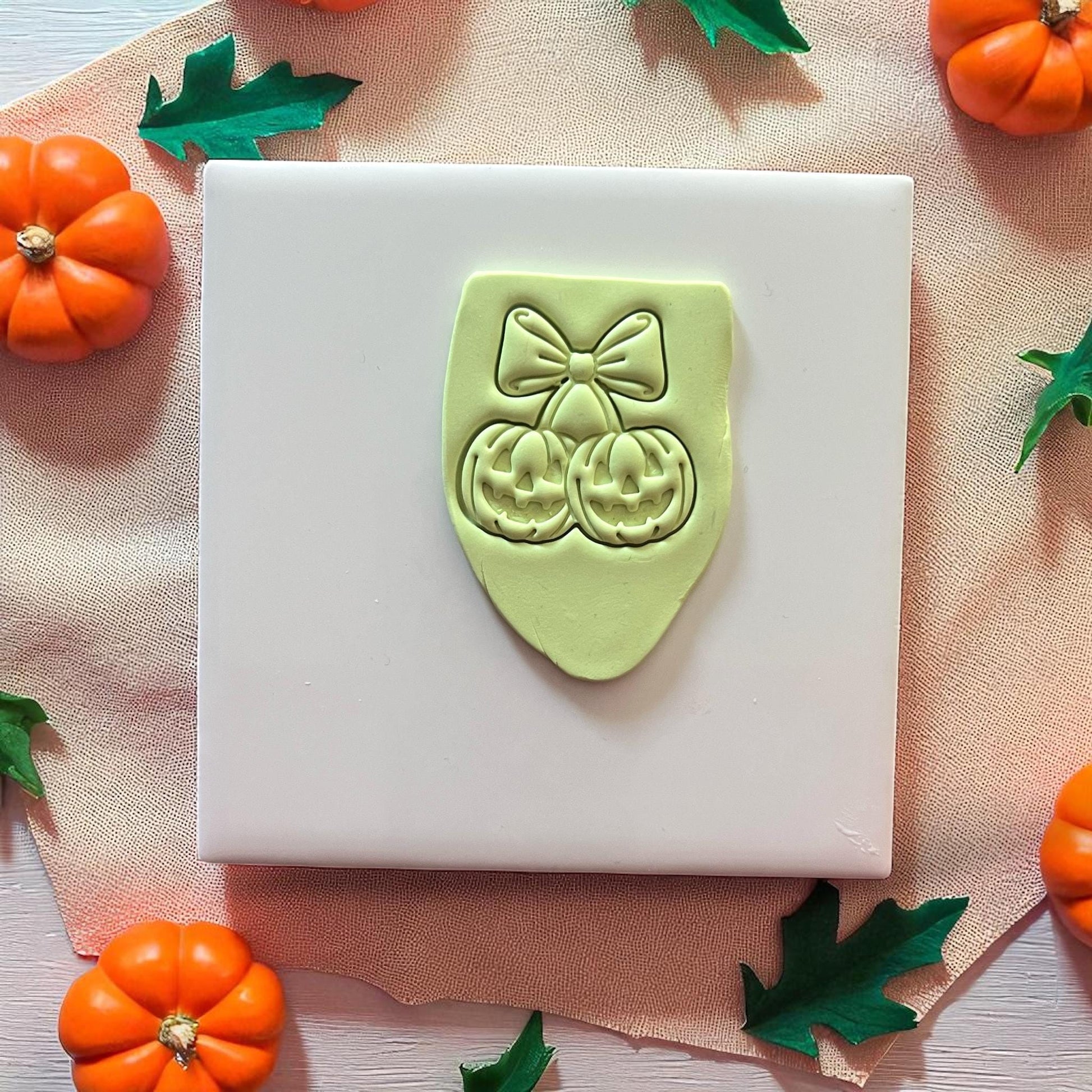 Green pumpkin cherry bow clay imprint on white tile with orange and green leaves.