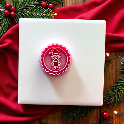 Pink scalloped circle elf clay cutter on white tile with red fabric and pine branches.
