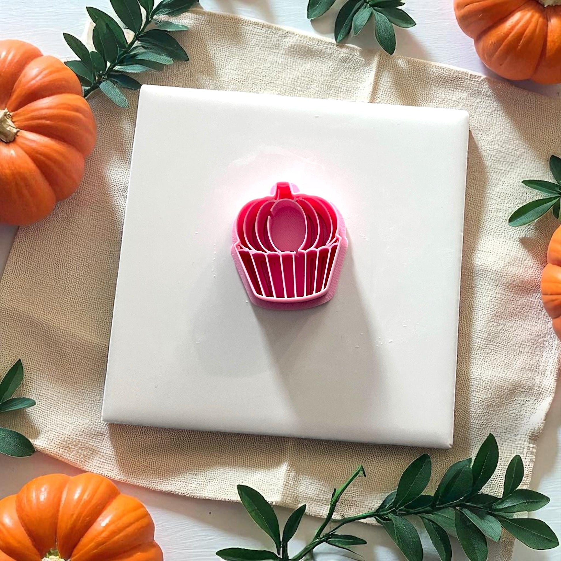 Pink pumpkin cupcake cutter on white tile surrounded by greenery