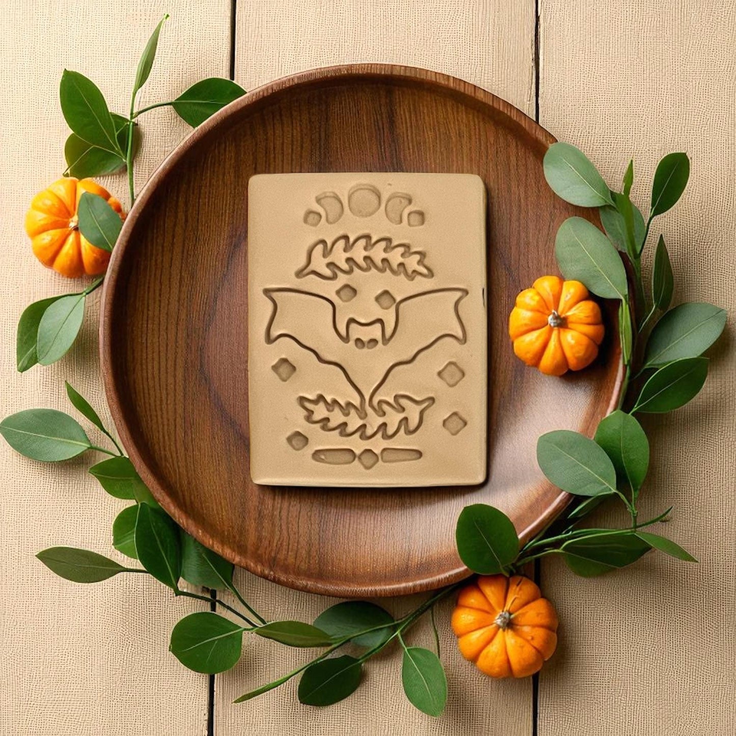 Large beige bat rectangle clay imprint on wooden plate with leaves and pumpkins.