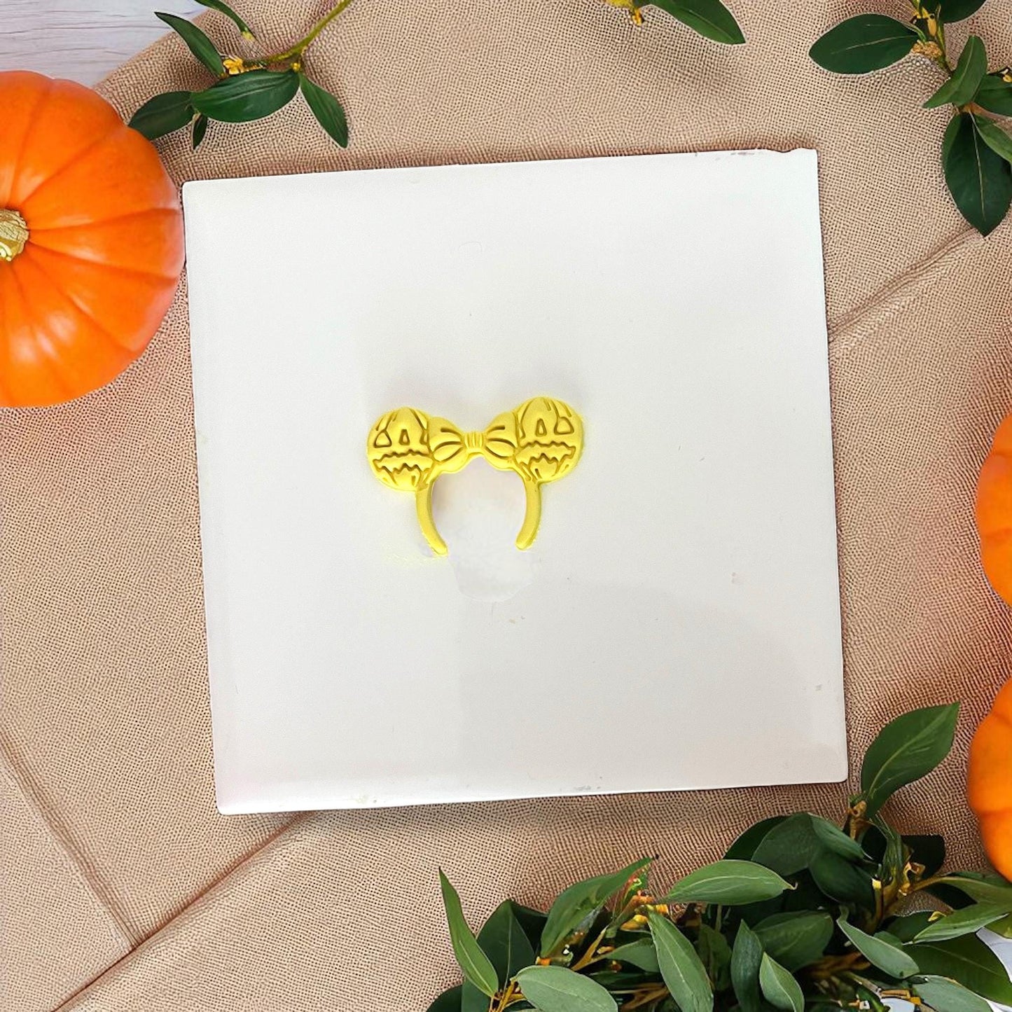 Yellow pumpkin bow ears clay imprint on white tile with greenery and pumpkins.