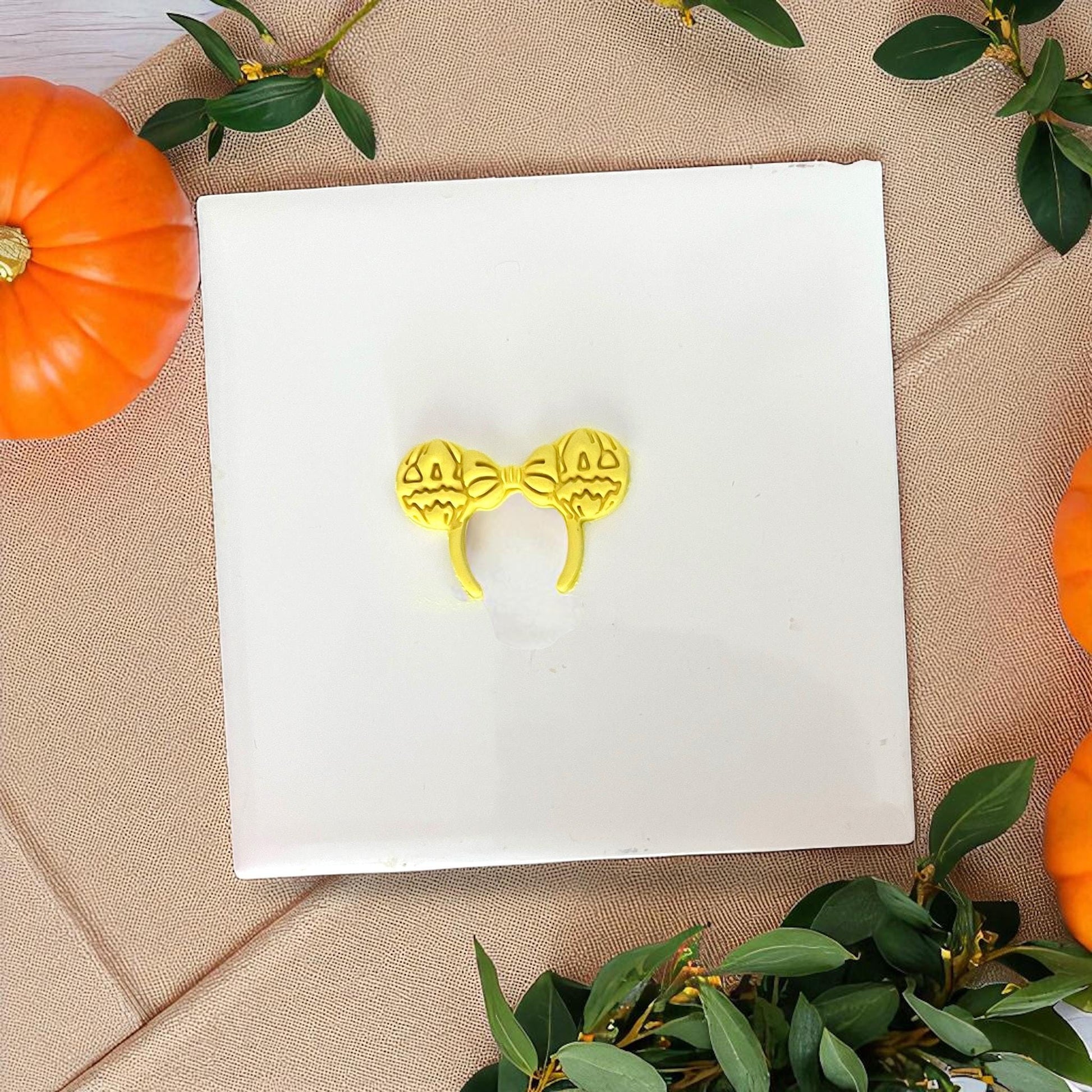 Yellow pumpkin bow ears clay imprint on white tile with greenery and pumpkins.