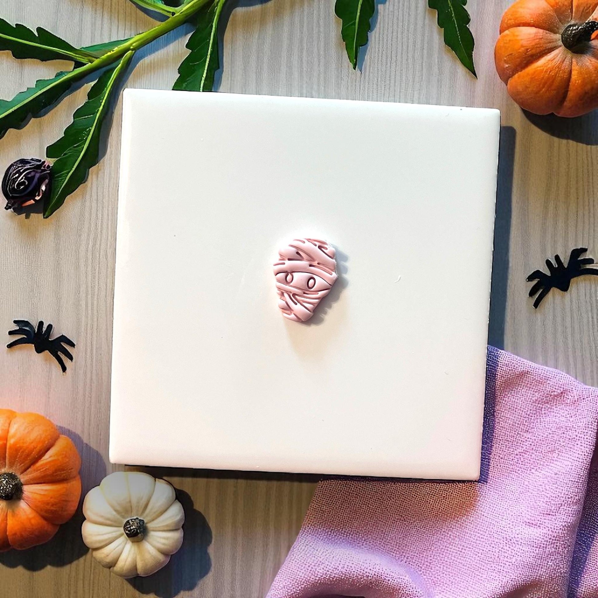 Small pink mummy clay piece on white tile with pumpkins and spiders