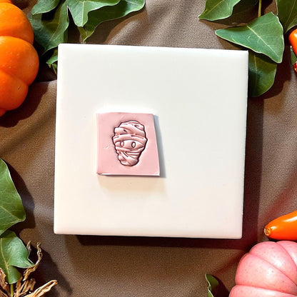 Pink mummy clay impression on square slab with pumpkins and greenery