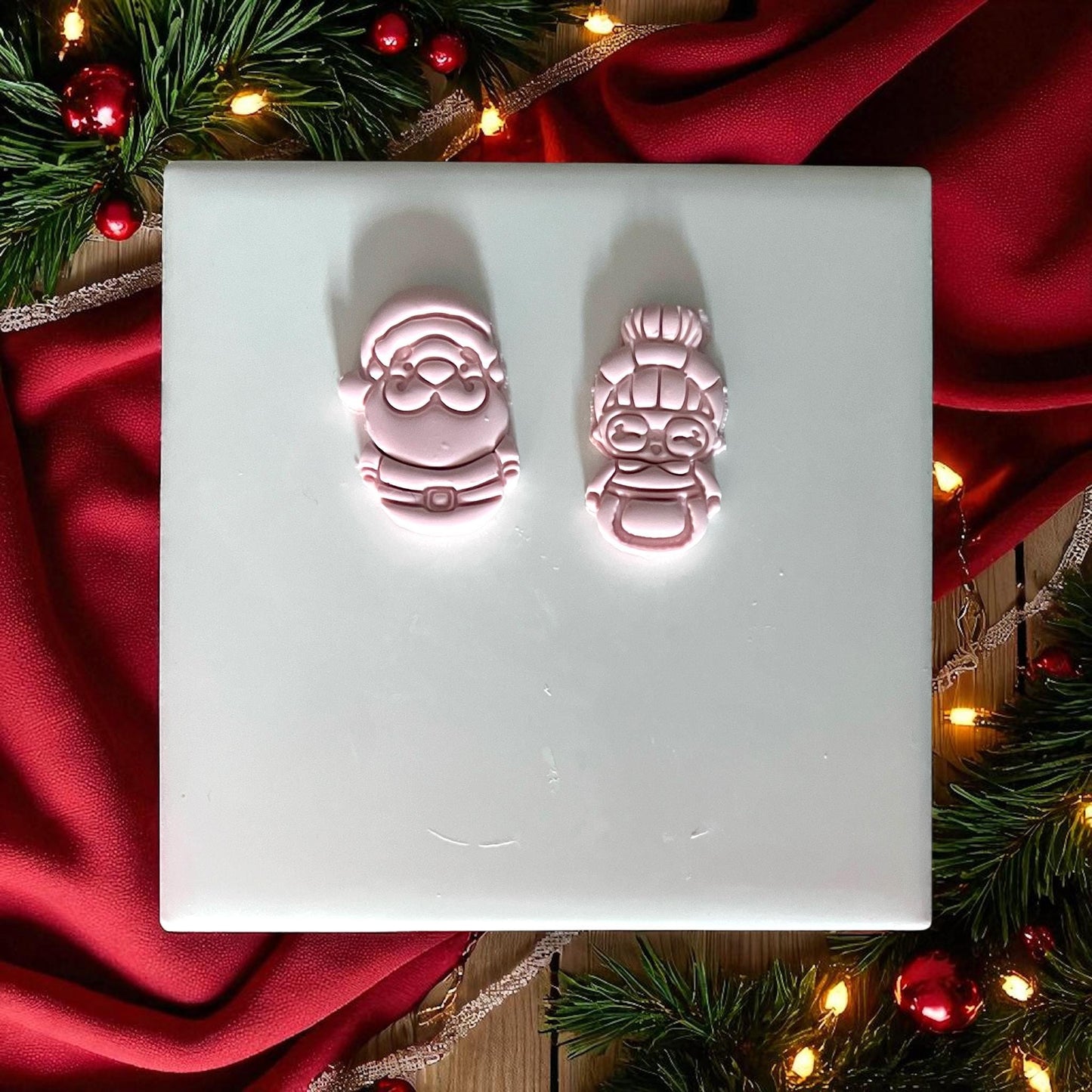 Pink clay Santa and Mrs Claus cutouts displayed on white tile with red fabric and pine branches