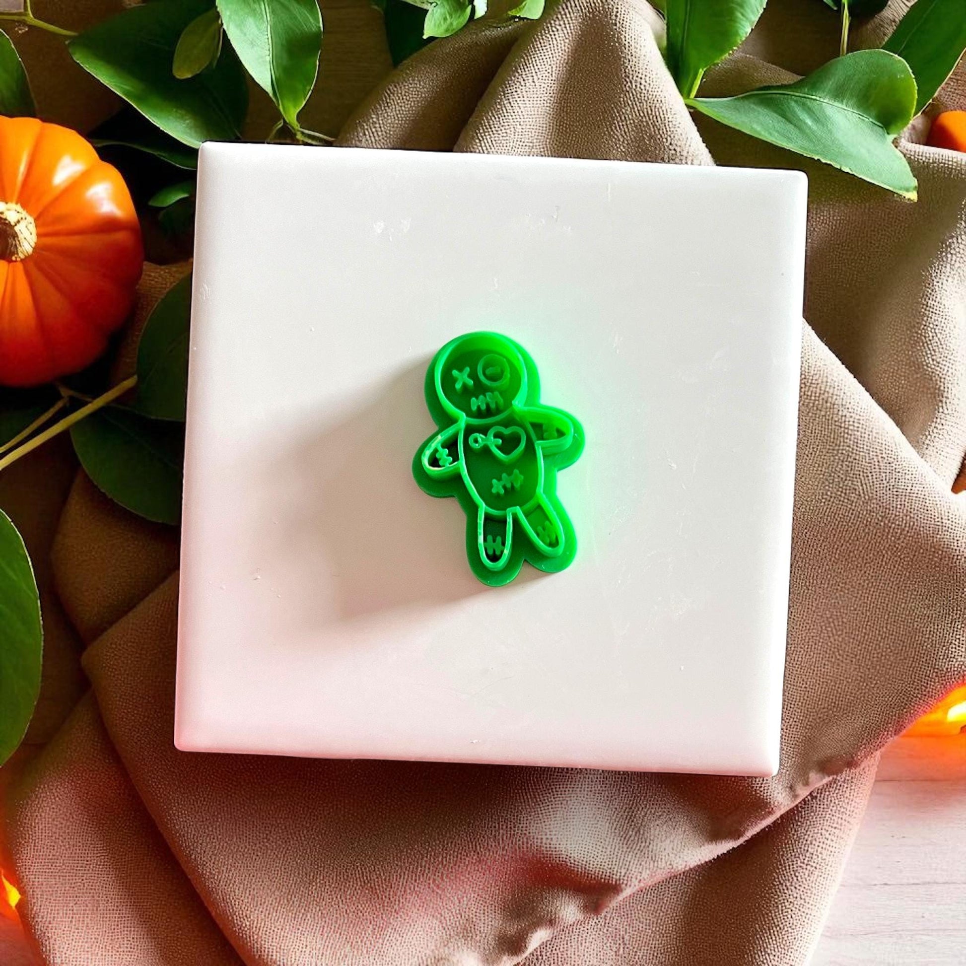Green voodoo doll clay cutter on white tile with pumpkins and greenery