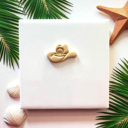 Beige cowboy hat clay piece with seashell detail on white tile with palm leaves