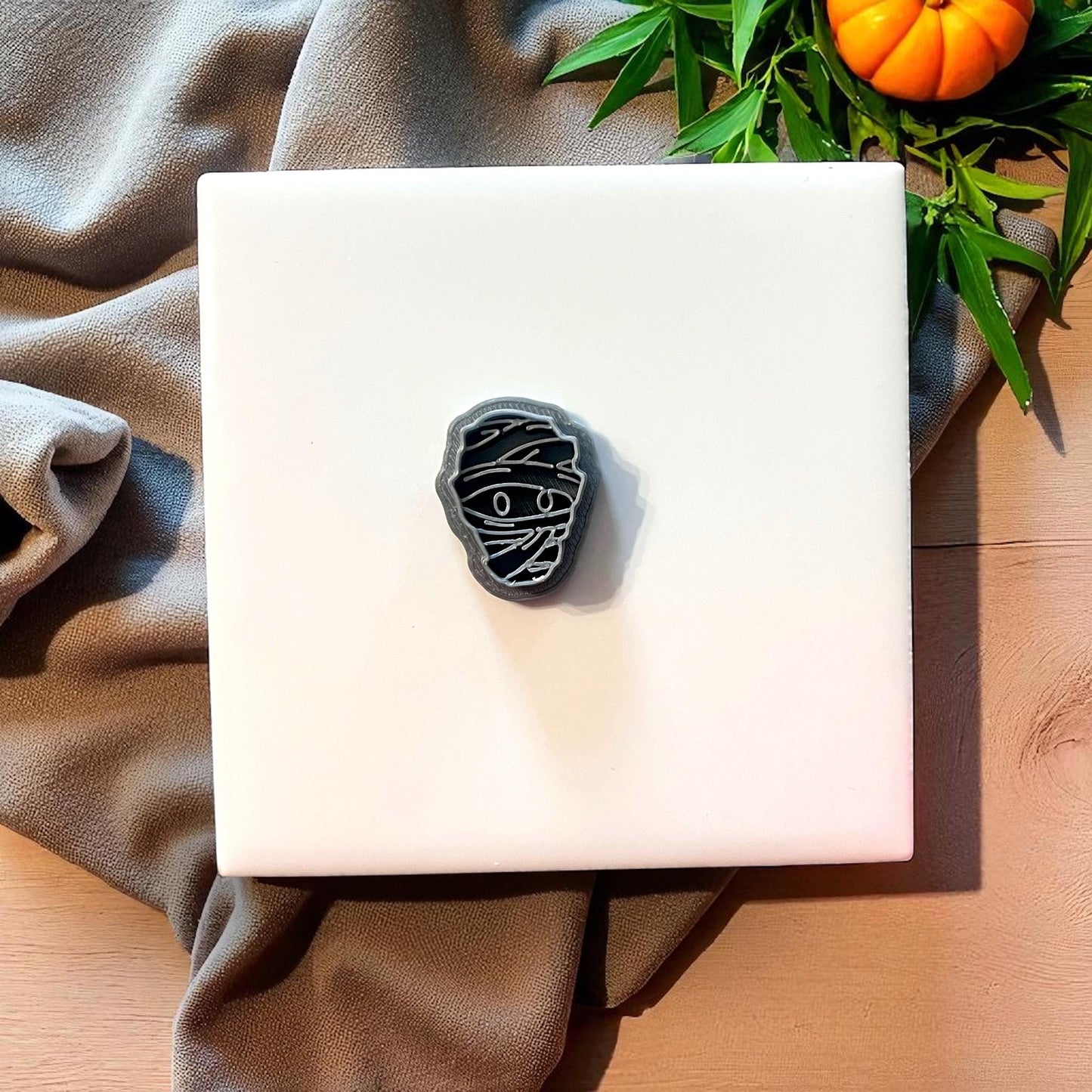 Black mummy face clay cutter on white tile with greenery and pumpkin