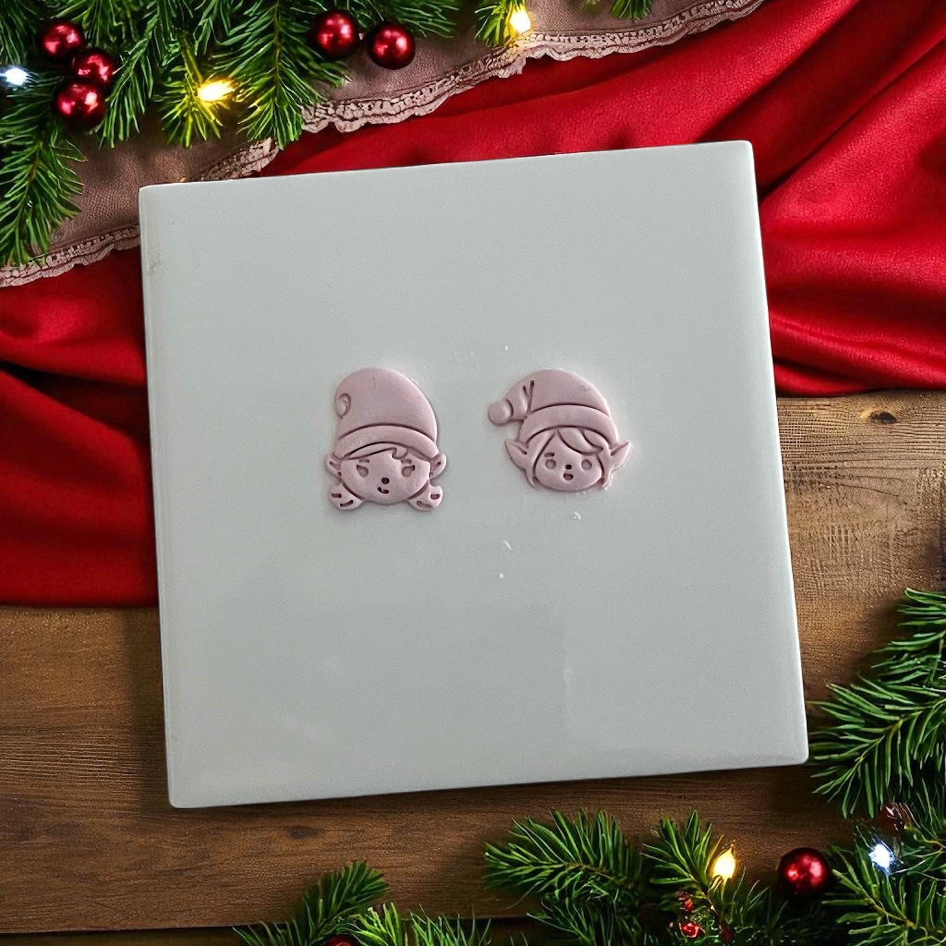 Elf boy and girl clay imprints on white tile with festive greenery and red fabric