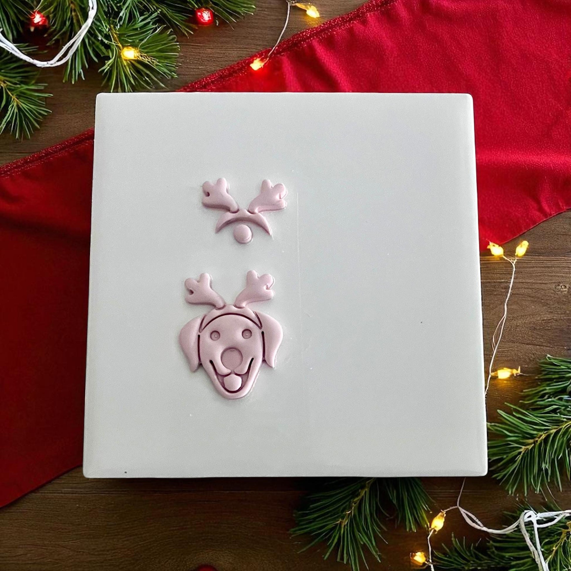 Clay imprint of labrador dog with reindeer ears and nose on white tile surrounded by Christmas lights