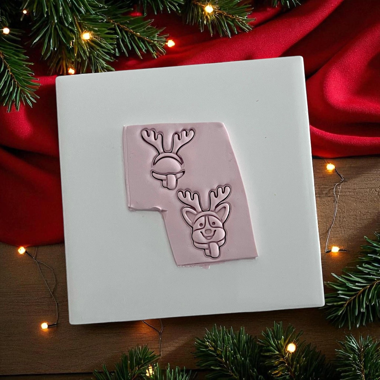 Pink clay imprint of corgi dog with reindeer antlers and tongue out on white tile with red cloth