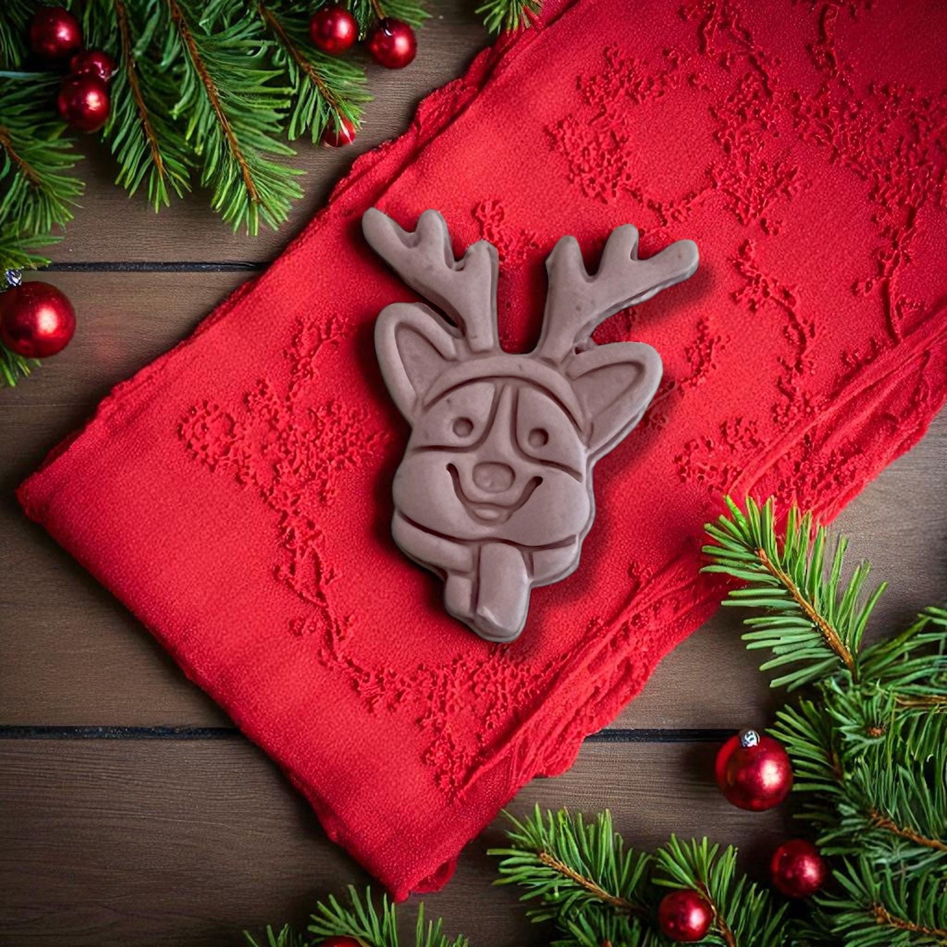 Brown clay imprint of corgi dog with reindeer ears and tongue on red cloth with Christmas greenery