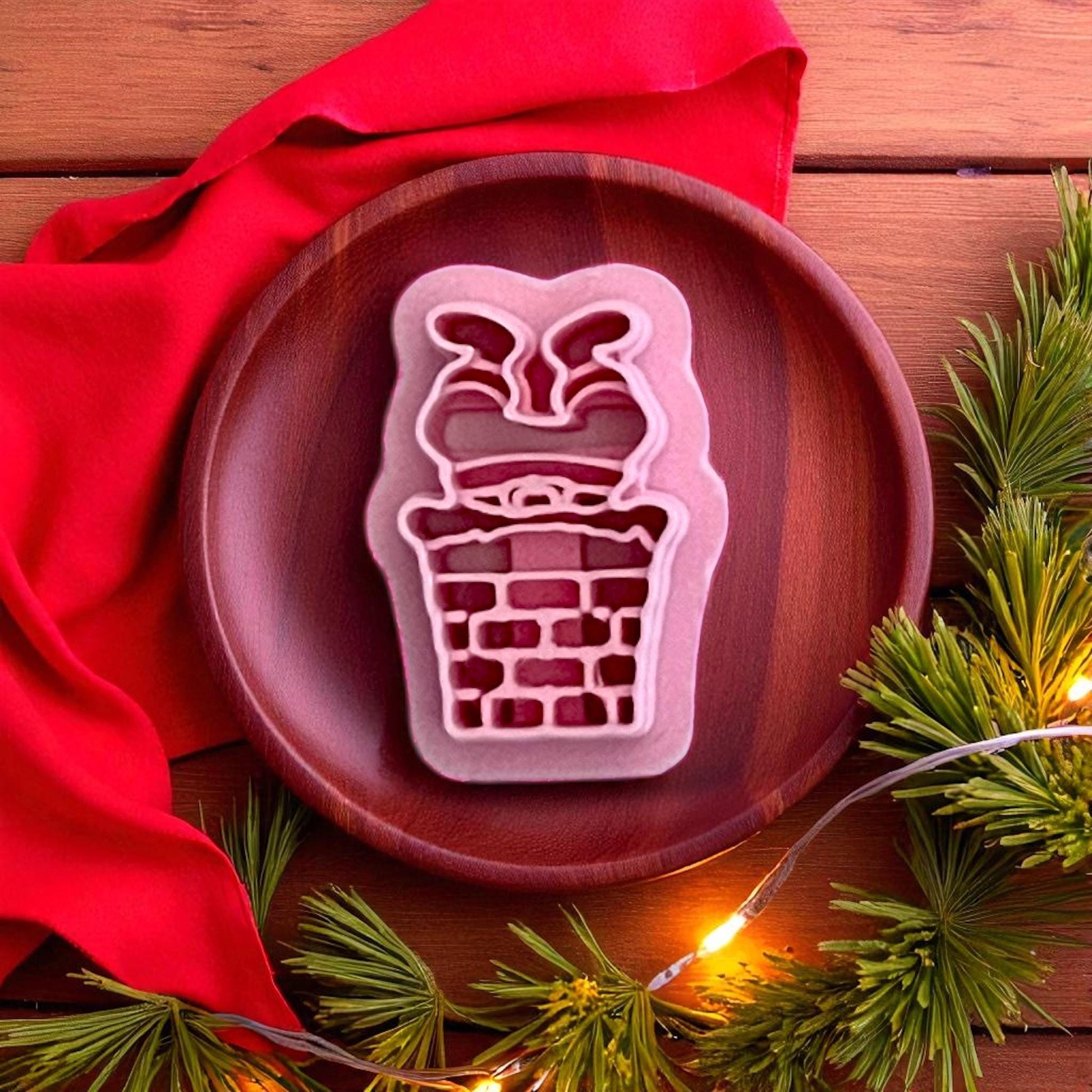 Pink polymer clay cutter shaped like Santa stuck in chimney on wooden plate with greenery