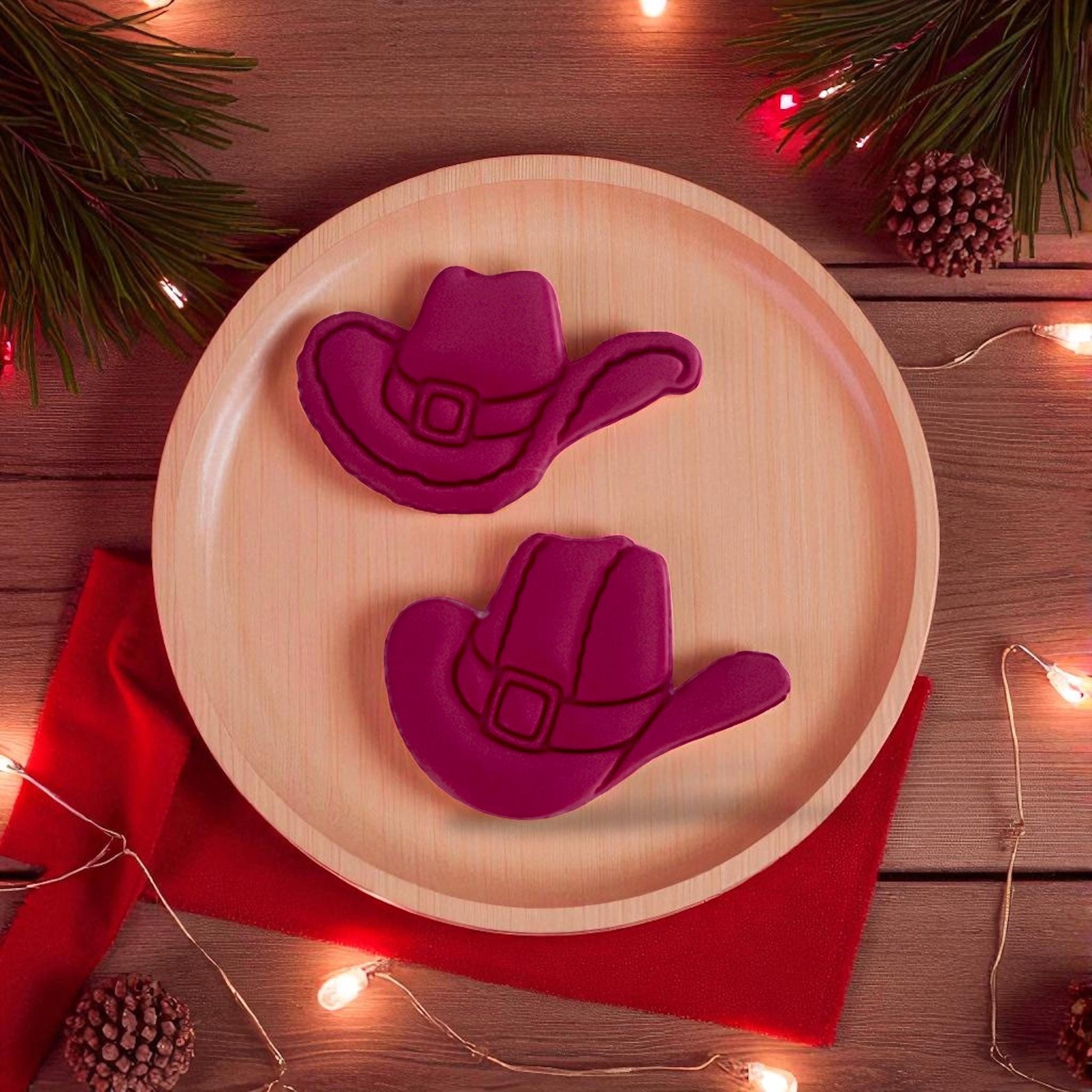 Two red clay cowboy hats displayed on wooden plate with holiday lights