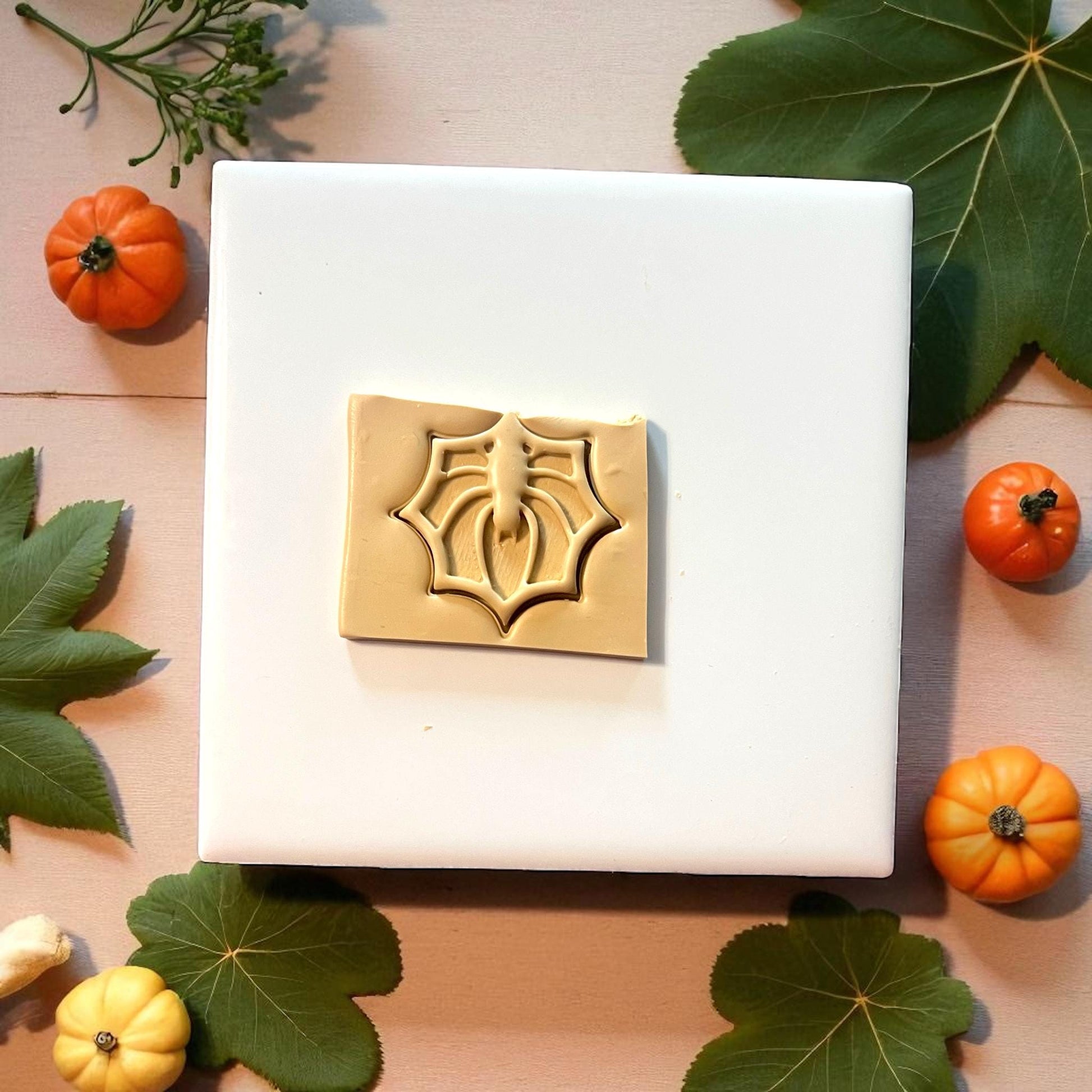 Beige spider web clay imprint on white tile with green leaves and pumpkins