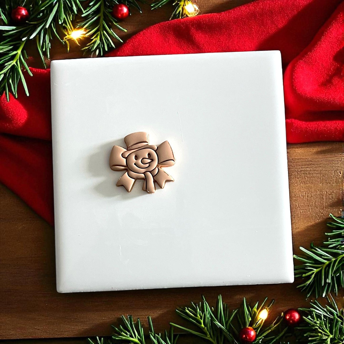 Polymer clay snowman impression with scarf and hat on white tile surrounded by pine branches