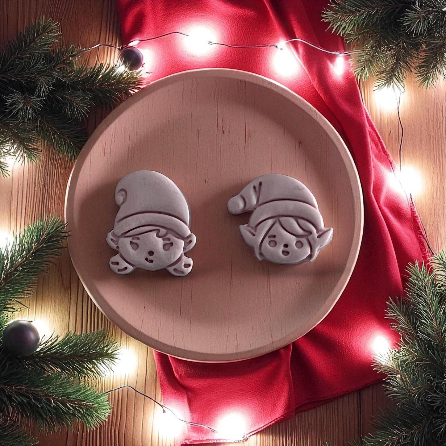 Clay imprints of elf boy and girl faces on wooden plate with red fabric and Christmas lights