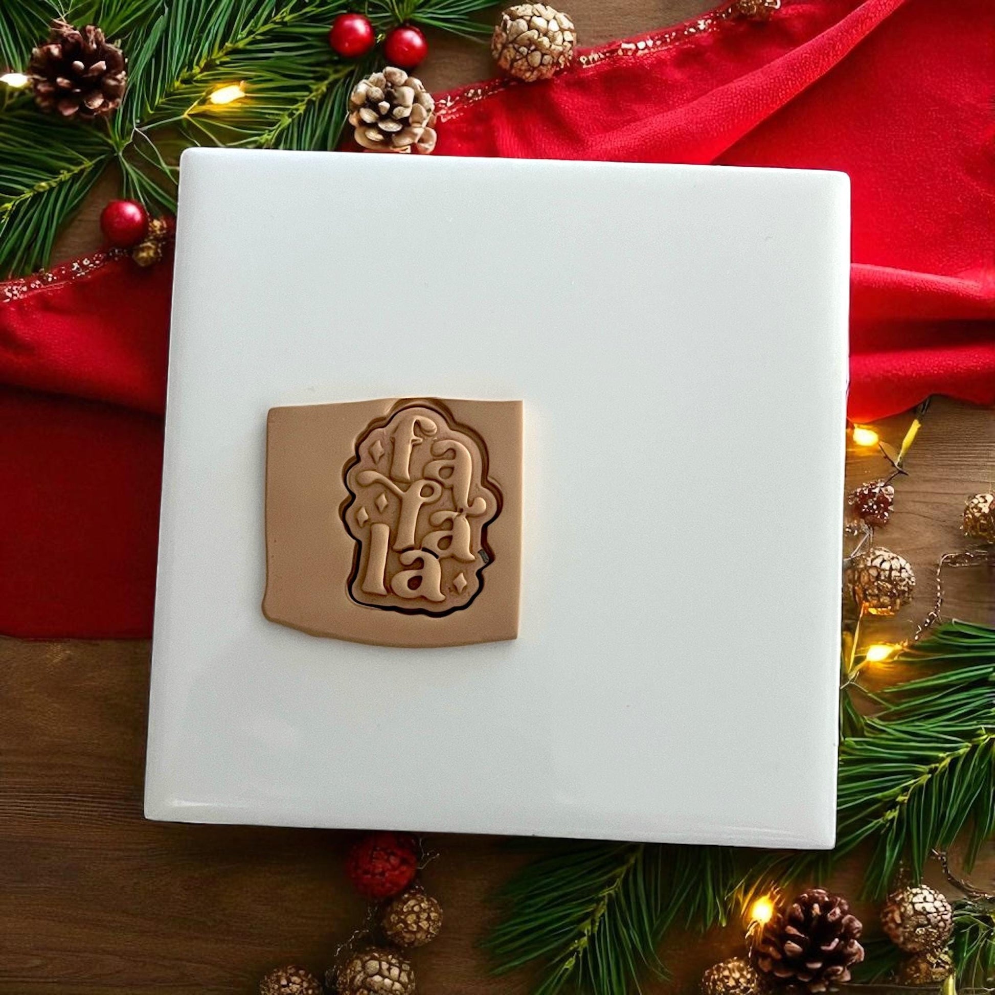 Festive Fa La La polymer clay piece on red fabric surrounded by pine branches and ornaments