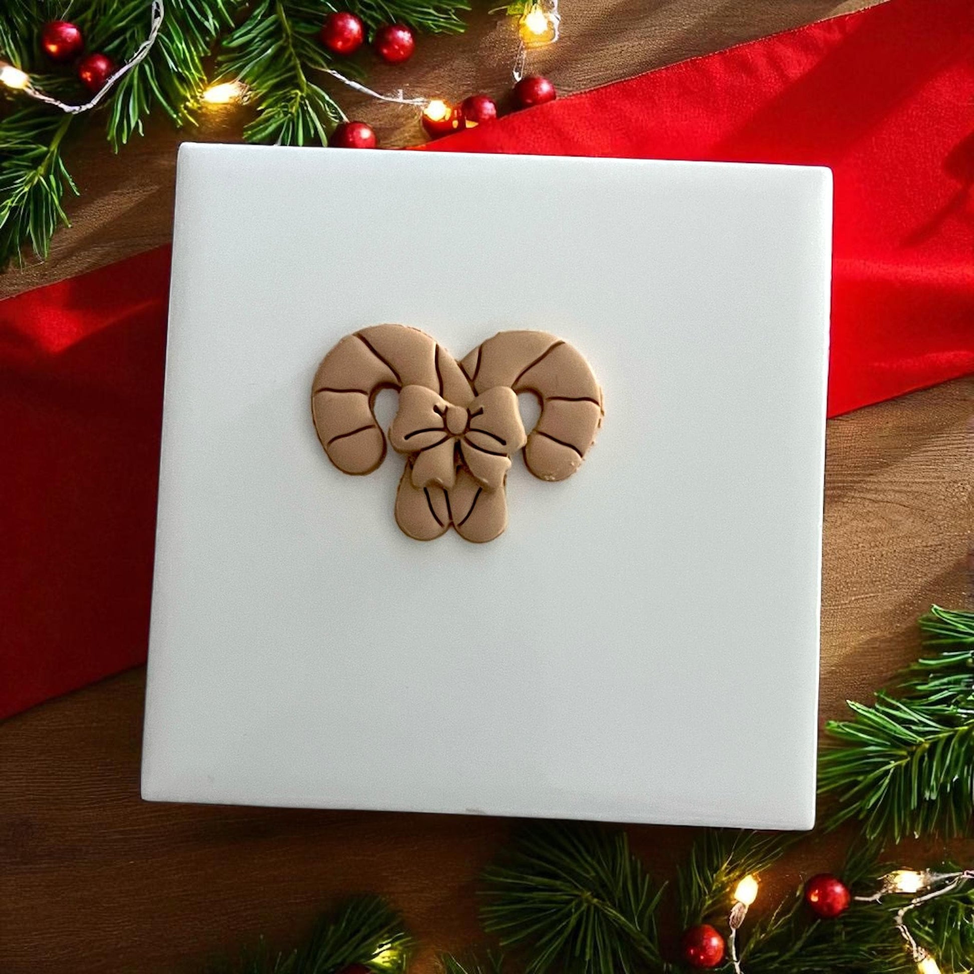 Candy cane bow clay design on white tile surrounded by Christmas lights and greenery