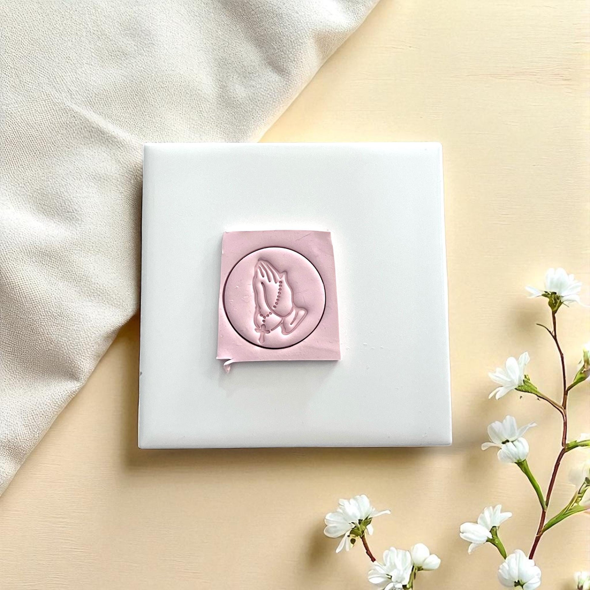 Large pink clay imprint of praying hands with rosary on soft beige fabric and round wooden plate