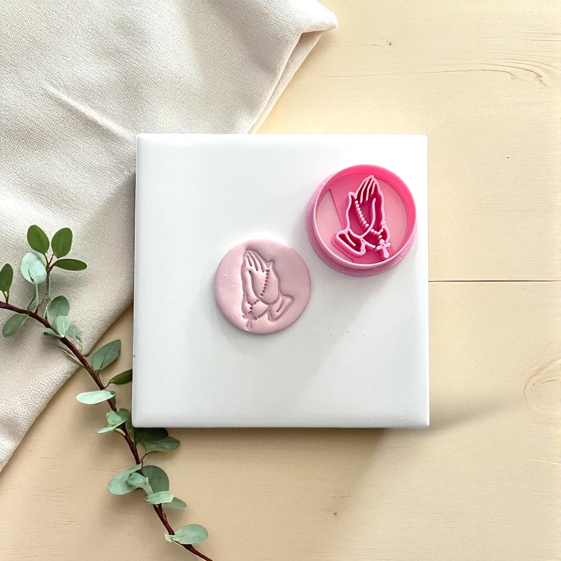 Pink cutter and clay imprint of praying hands with rosary side by side on white tile