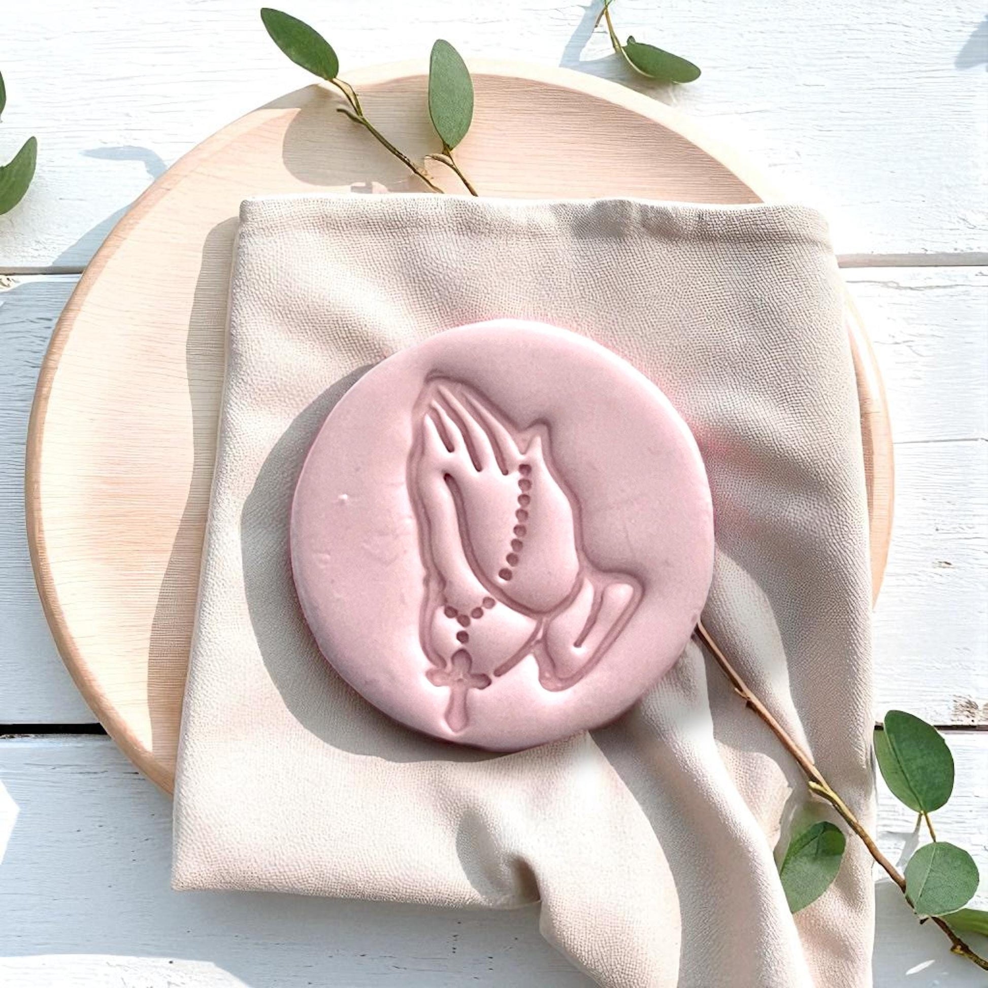 Large pink clay imprint of praying hands with rosary on soft beige fabric and round wooden plate