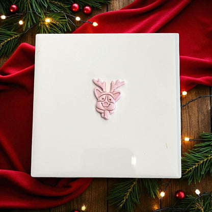 Pink clay imprint of corgi face with reindeer ears and tongue on white tile with greenery