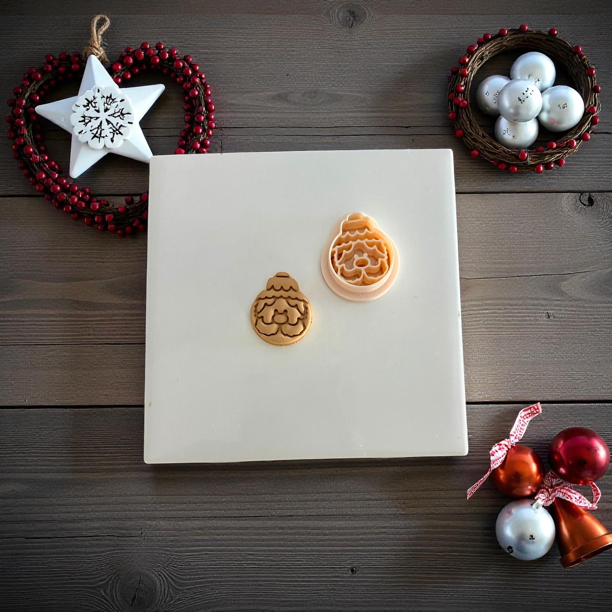 Two Santa ornament clay imprints side by side on white tile surrounded by ornaments