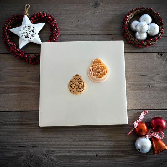Two Santa ornament clay imprints side by side on white tile surrounded by ornaments