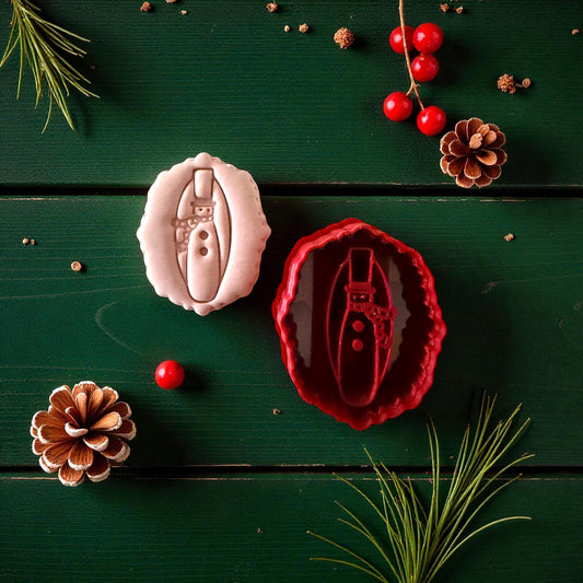 Pink snowman clay imprint beside red cutter on green wood with pinecones and berries