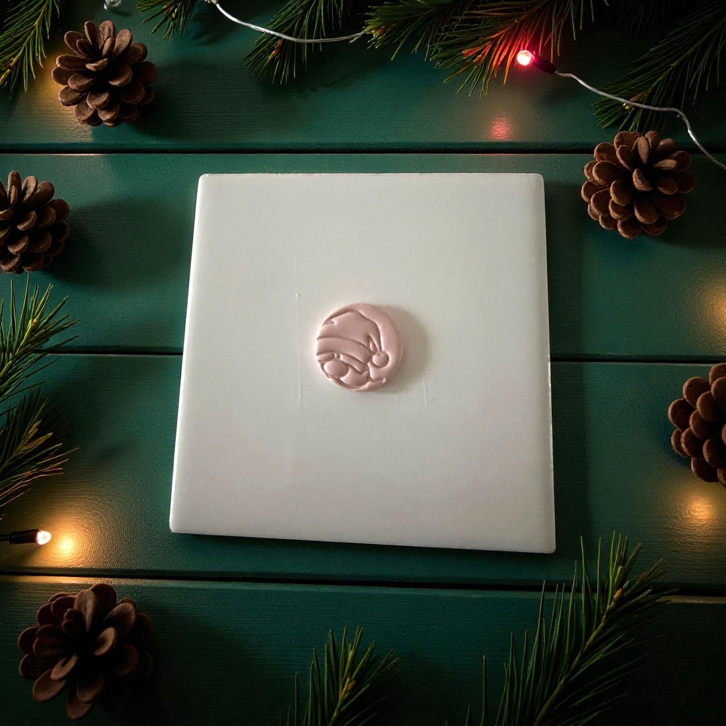 Pink peeking Santa clay imprint centered on white tile with pine and holiday lights