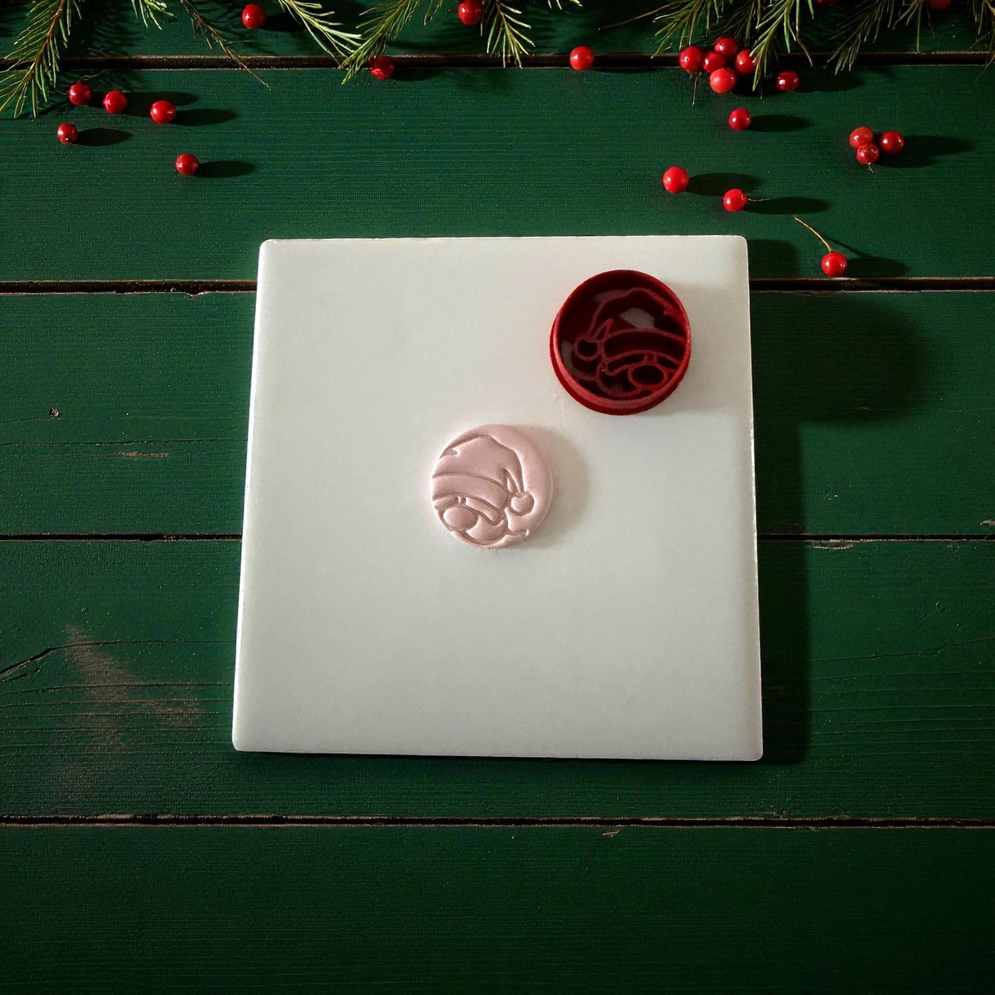 Pink Santa clay imprint beside red round cutter on white tile with holly and berries