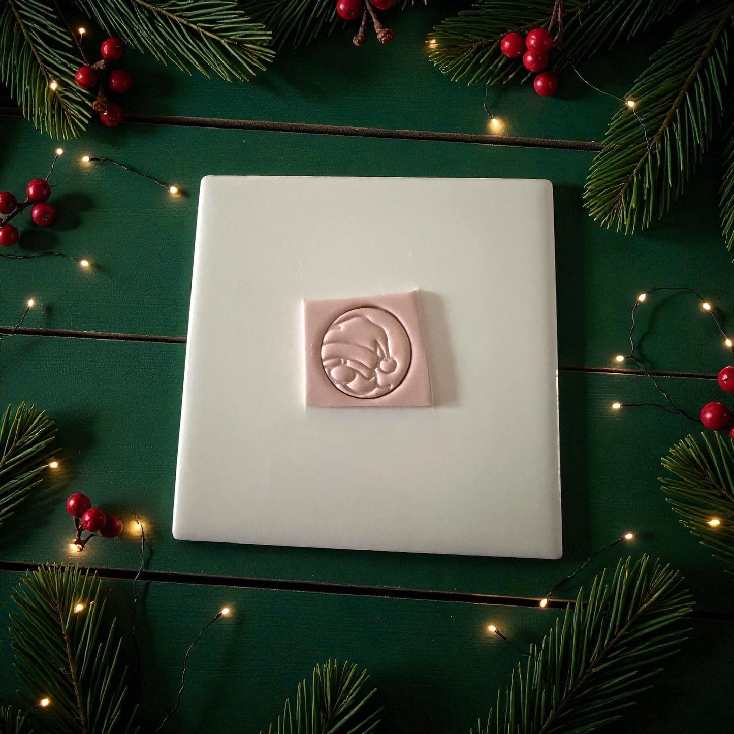 Pink peeking Santa clay imprint centered on white tile with pine and holiday lights