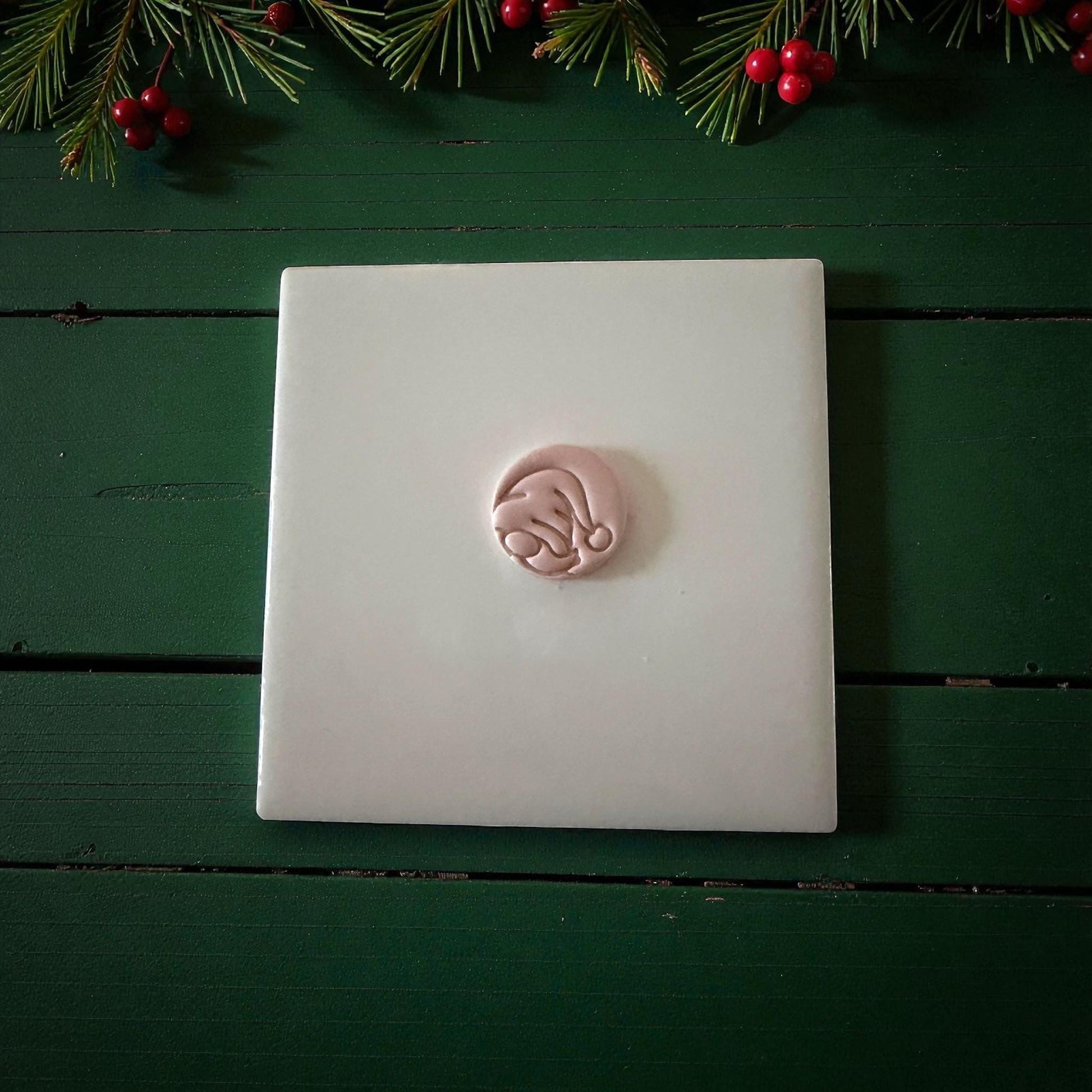 Small round gnome clay imprint centered on white tile with Christmas greenery