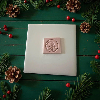 Pink peeking gnome clay imprint on white tile surrounded by pinecones and berries