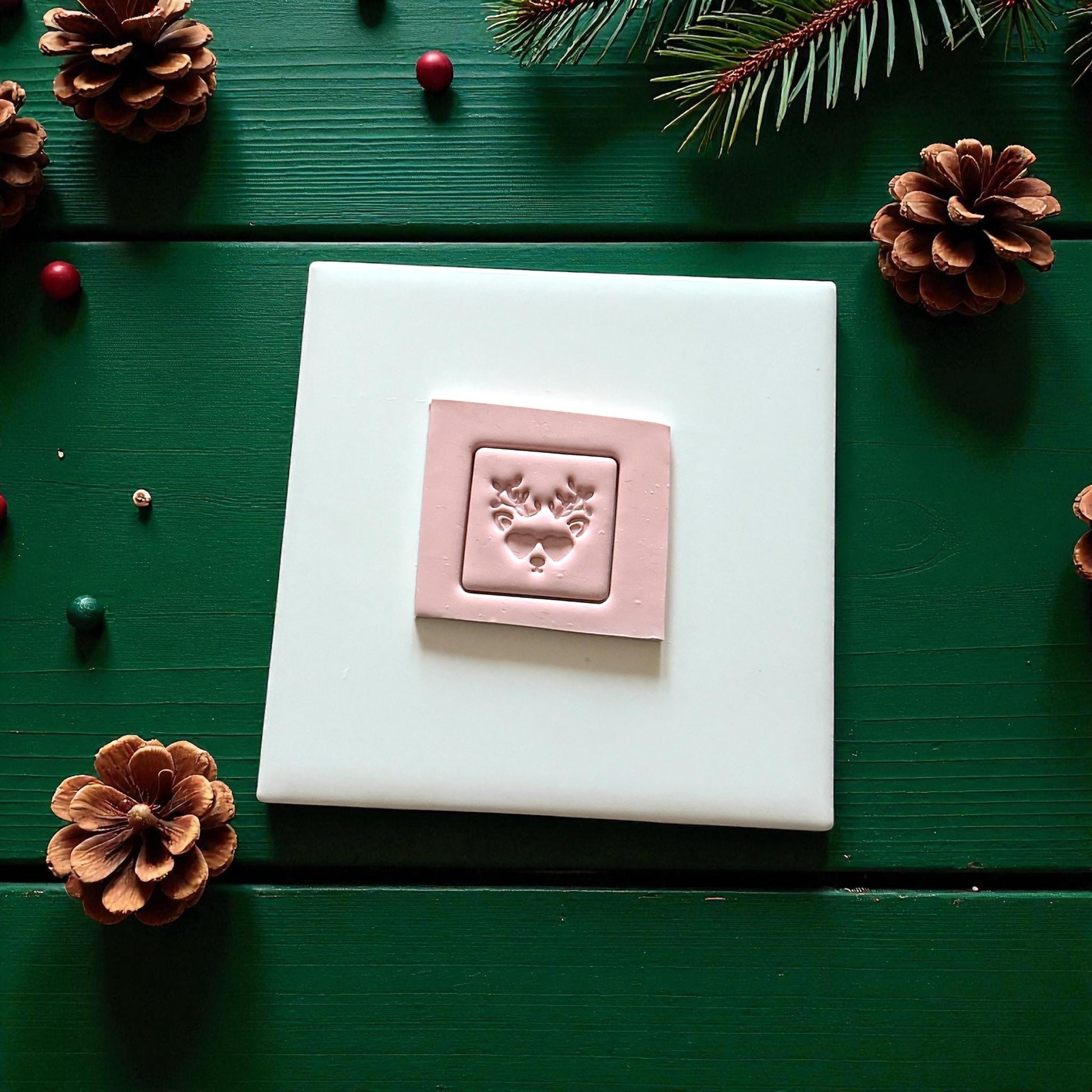 Pink reindeer face clay imprint on white tile with pinecones and berries