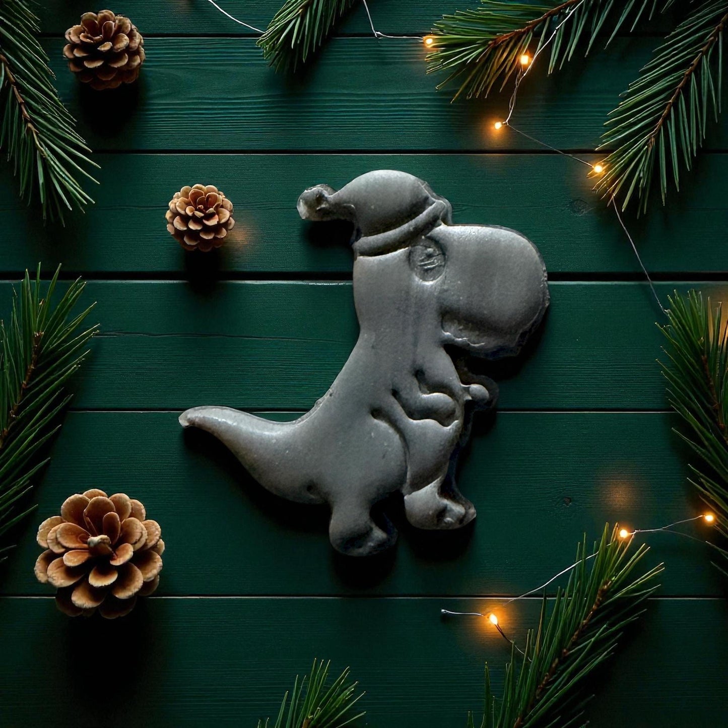 Close-up of gray dino clay imprint wearing Santa hat surrounded by pinecones and lights