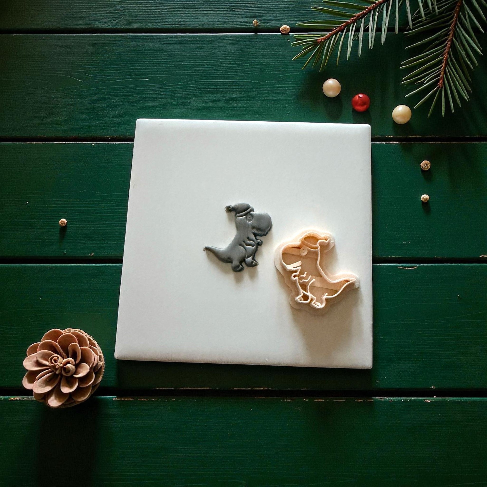 Dino clay imprint beside light-colored cutter on white tile with pinecone and berries