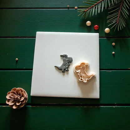 Dino clay imprint beside light-colored cutter on white tile with pinecone and berries