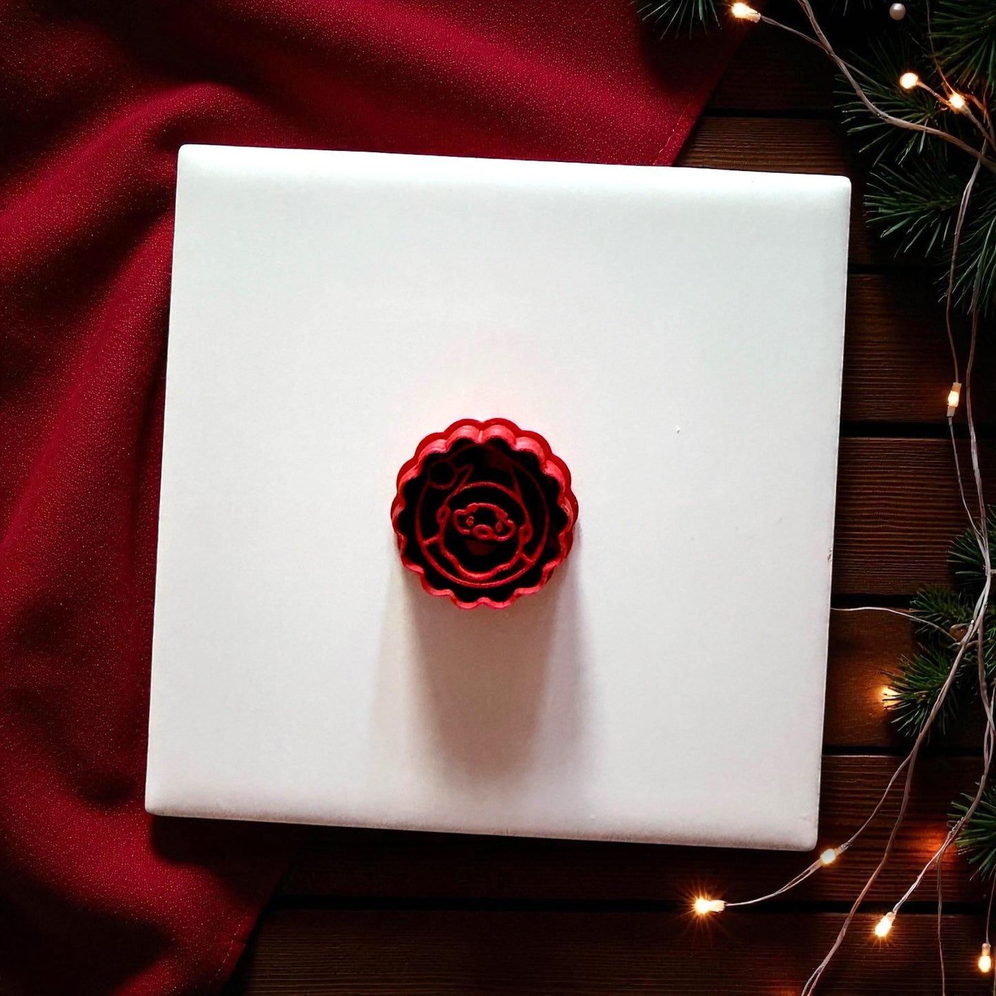 Small red scalloped Santa cutter centered on white tile with Christmas lights and red cloth