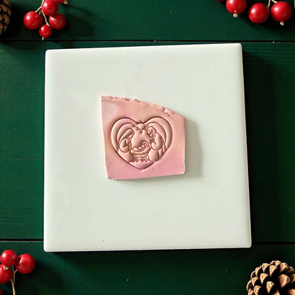 Pink nativity heart clay imprint on white tile surrounded by pinecones and red berries